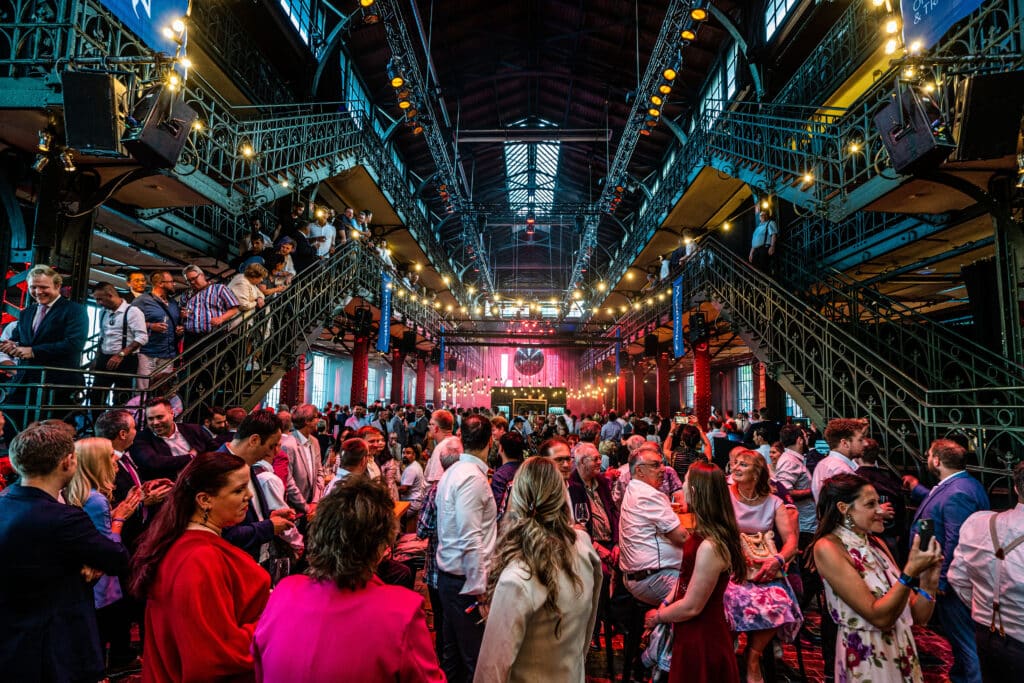 Gut besuchte Abendveranstaltung in einer historischen Industriehalle mit gusseisernen Treppen, Lichterketten und Bühne im Hintergrund