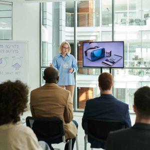 Rear view of young audience listening to mature female coach at seminar Rear view of young audience listening to mature female coach standing by interactive board with VR headset and smartphone at seminar